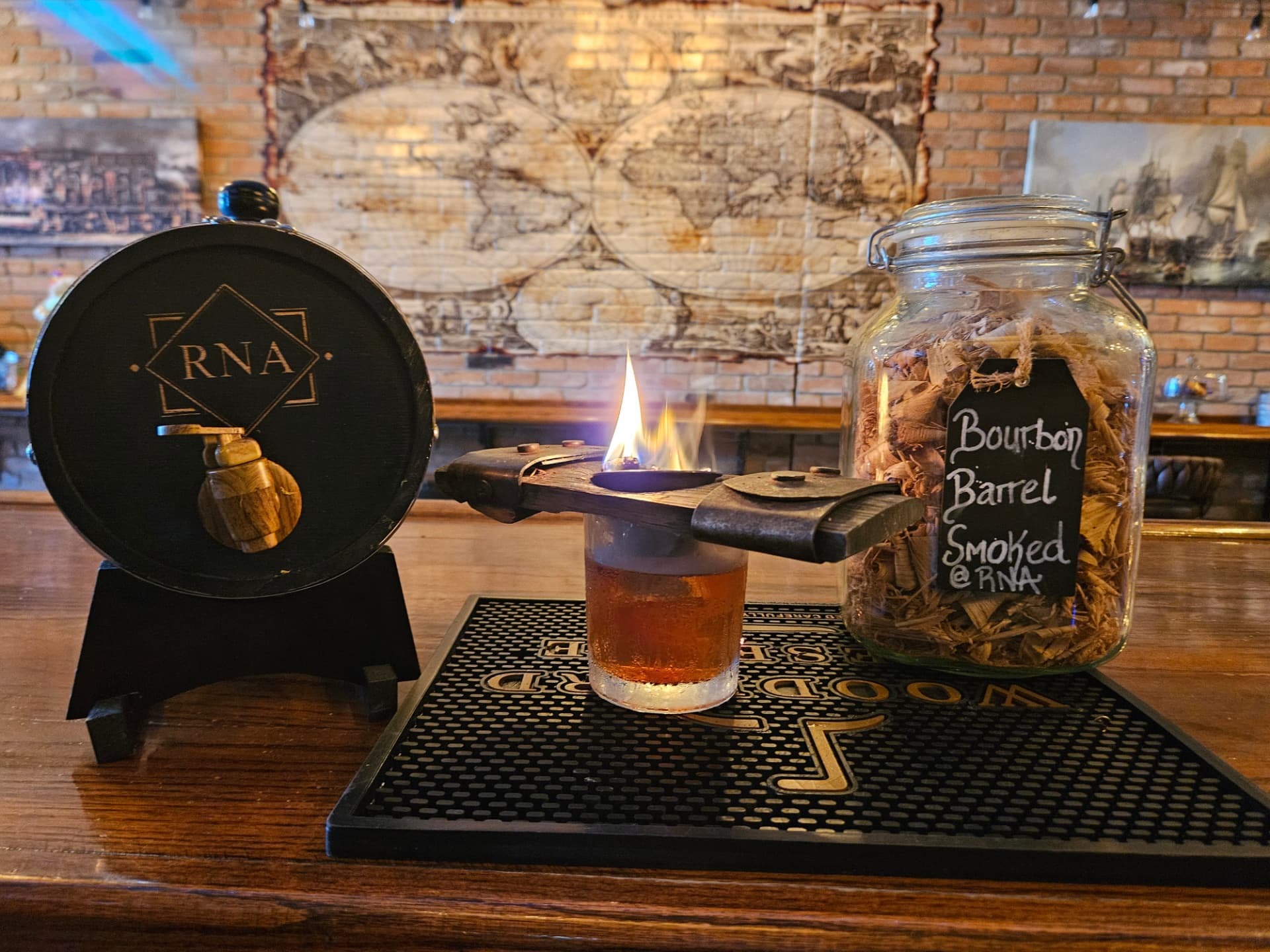 A bourbon barrel smoked cocktail flaming beside an RNA barrel and jar of wood chips at RNA Tavern